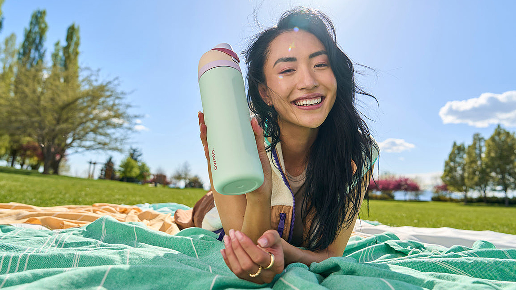 Woman holding mint green water bottle with burgundy trim on picnic blanket outdoors, smiling in sunny park setting