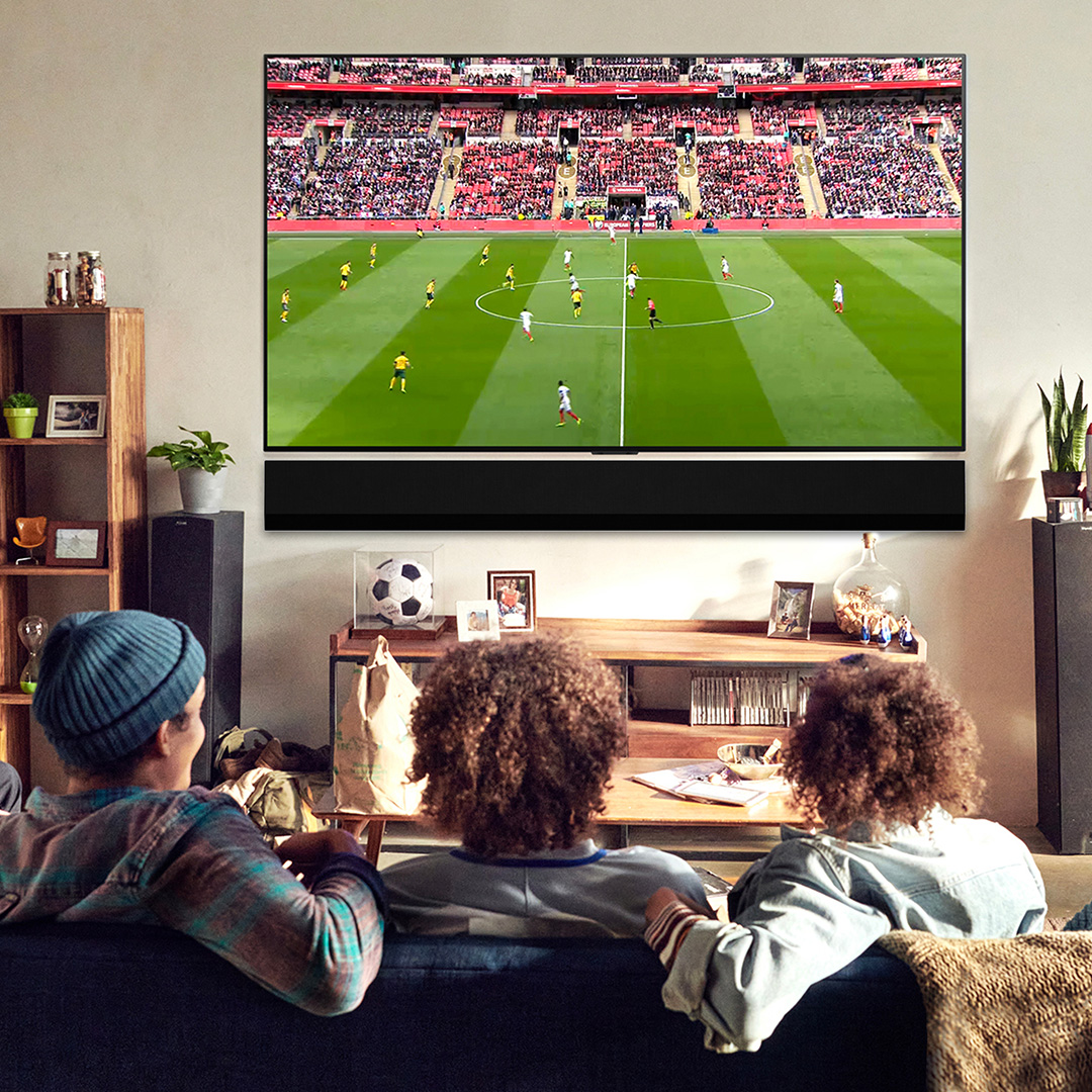 Group of people watching football match on large television screen in modern living room with wooden furniture and plants