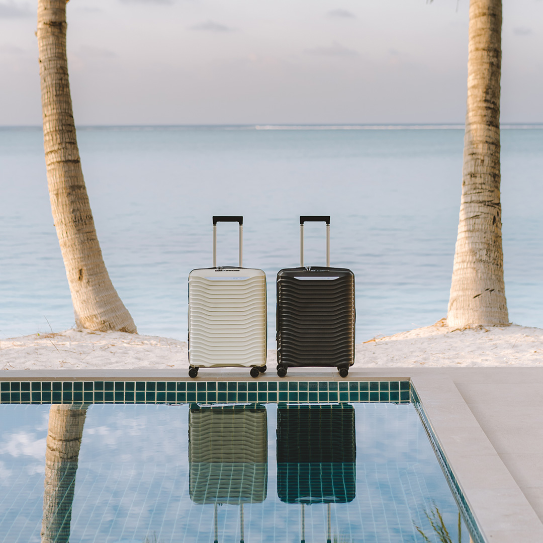 Cream and black ridged suitcases on a pool edge with palm trees and beach vista in background