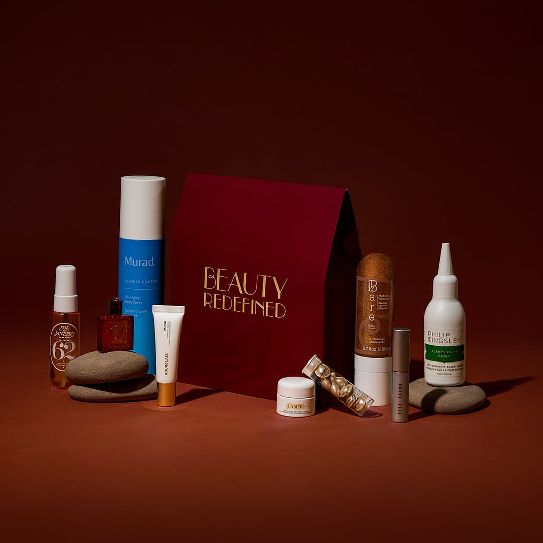 Assorted beauty products displayed against a dark red background. Items include creams, serums, and sprays from various brands, along with a red gift box labelled 'Beauty Redefined.' Stones are artistically placed among the products.