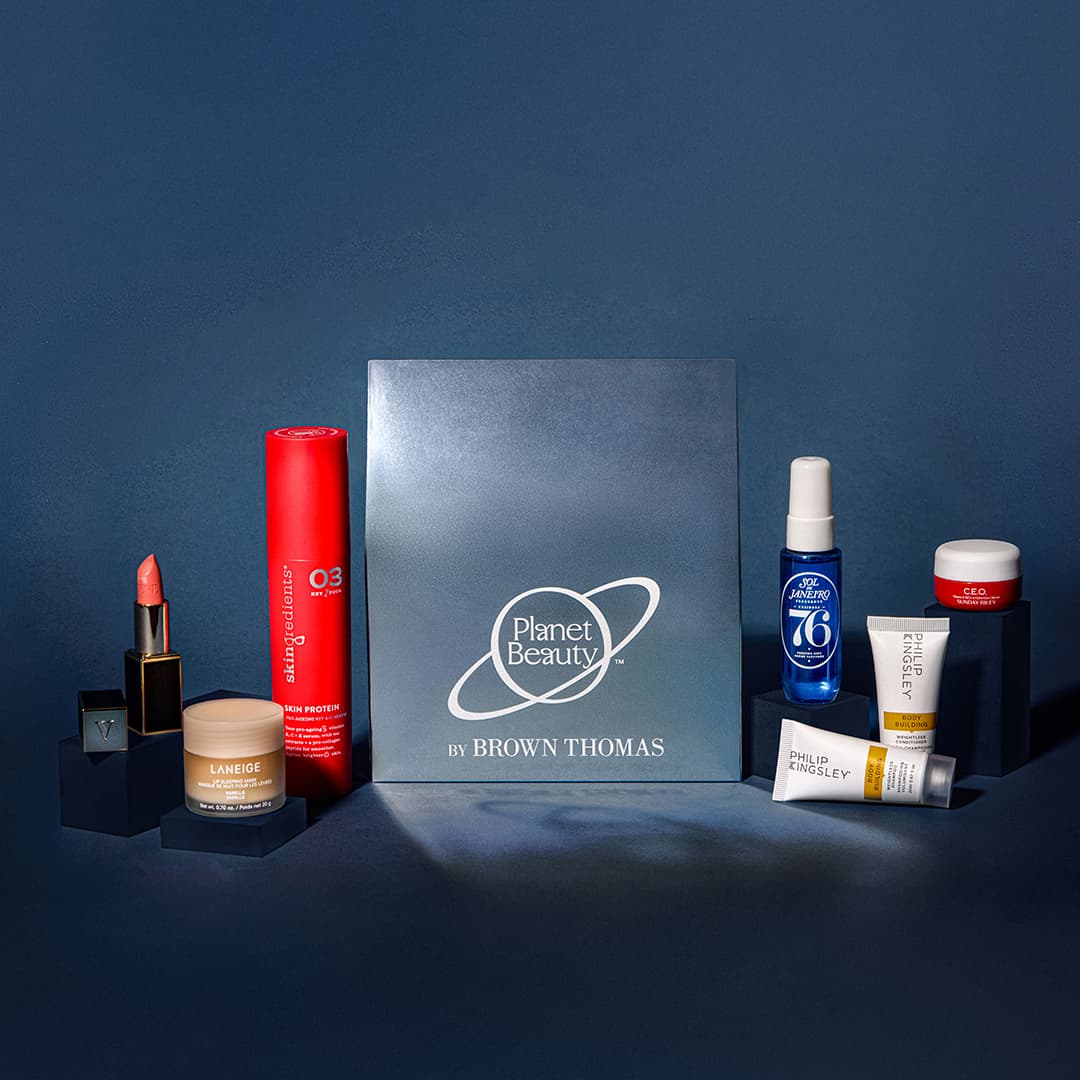 A collection of beauty products arranged around a 'Planet Beauty by Brown Thomas' box. Items include a red lipstick, moisturiser, red bottle of hair spray, blue facial toner, and various creams and skincare products, displayed against a dark blue background.