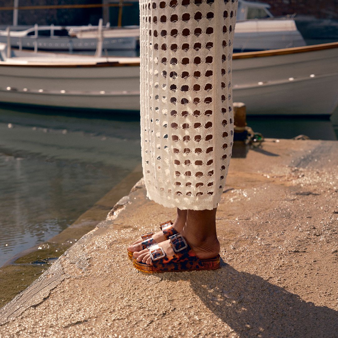 Person wearing orange patterned sandals standing on sandy beach near moored boats