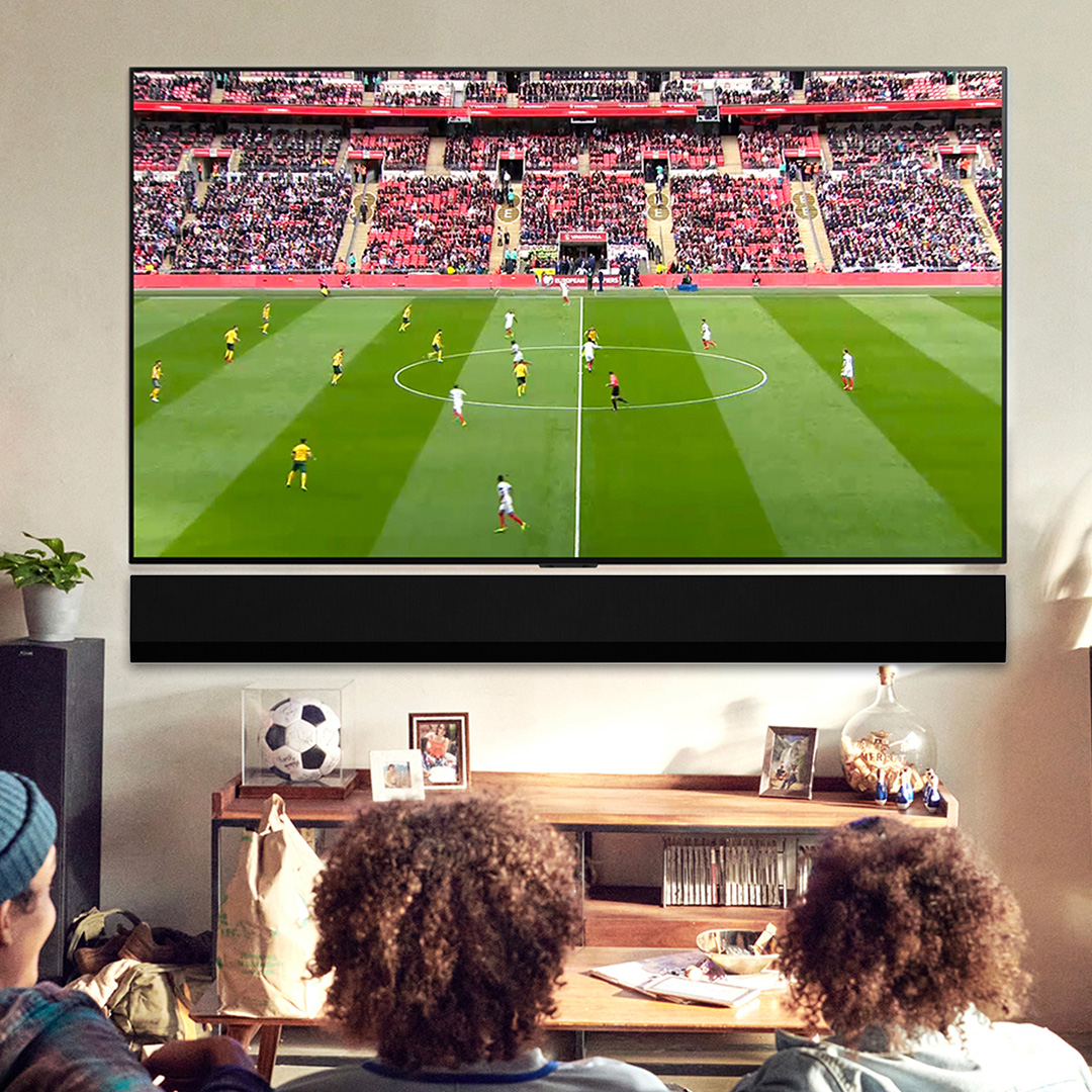 Three people sitting on sofa watching football match on large wall-mounted television, living room with wooden shelving and decor