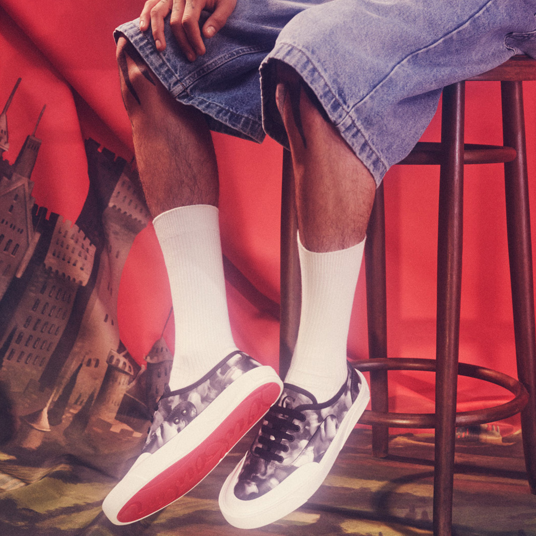 Person wearing white socks and patterned trainers with red sole detail, standing in front of urban cityscape backdrop with red lighting