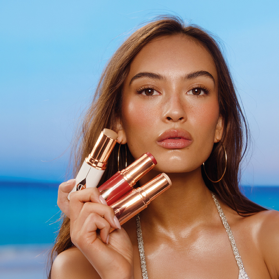 Woman holding Charlotte Tilbury highlighter and lipstick products against blue ocean background