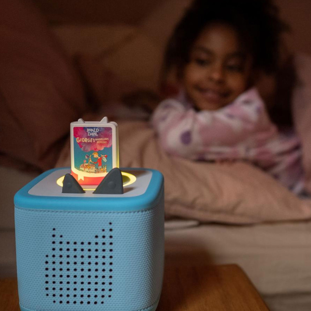 Blue speaker toy with book-shaped top displaying Roald Dahl George's Marvellous Medicine, child in background interacting with toy