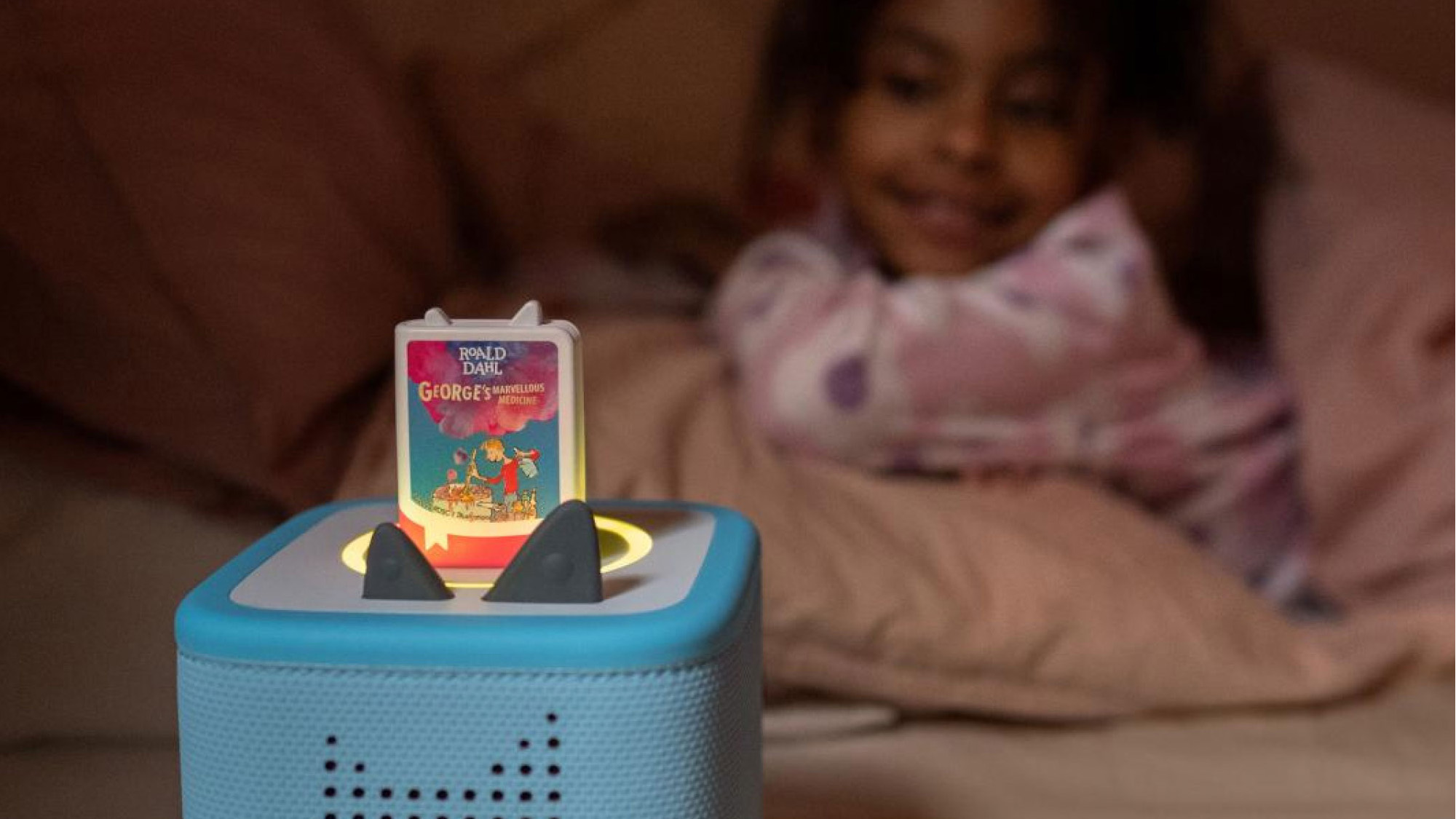 Blue speaker toy with book-shaped top displaying Roald Dahl George's Marvellous Medicine, child in background interacting with toy