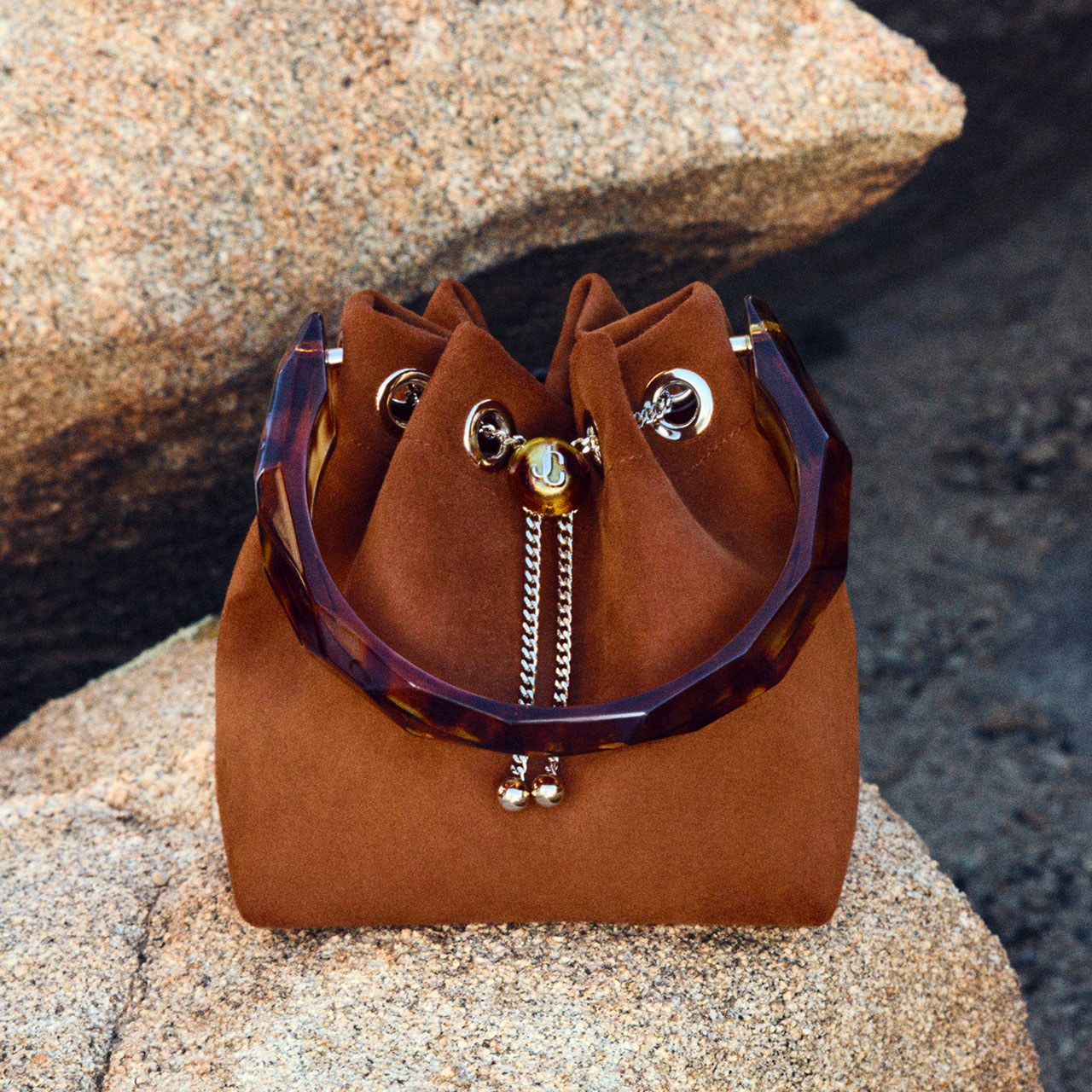 Rust-coloured suede bucket bag with tortoiseshell handles and silver chain drawstring on sandy beach