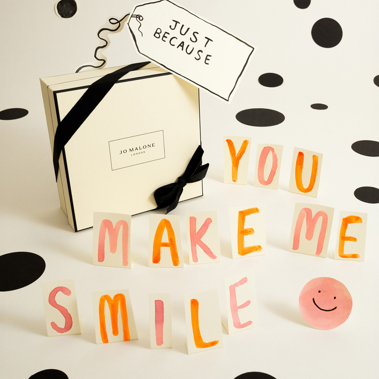 Jo Malone London fragrance gift box with black ribbon surrounded by colourful letter tiles spelling 'You Make Me Smile' and a smiley face decoration.