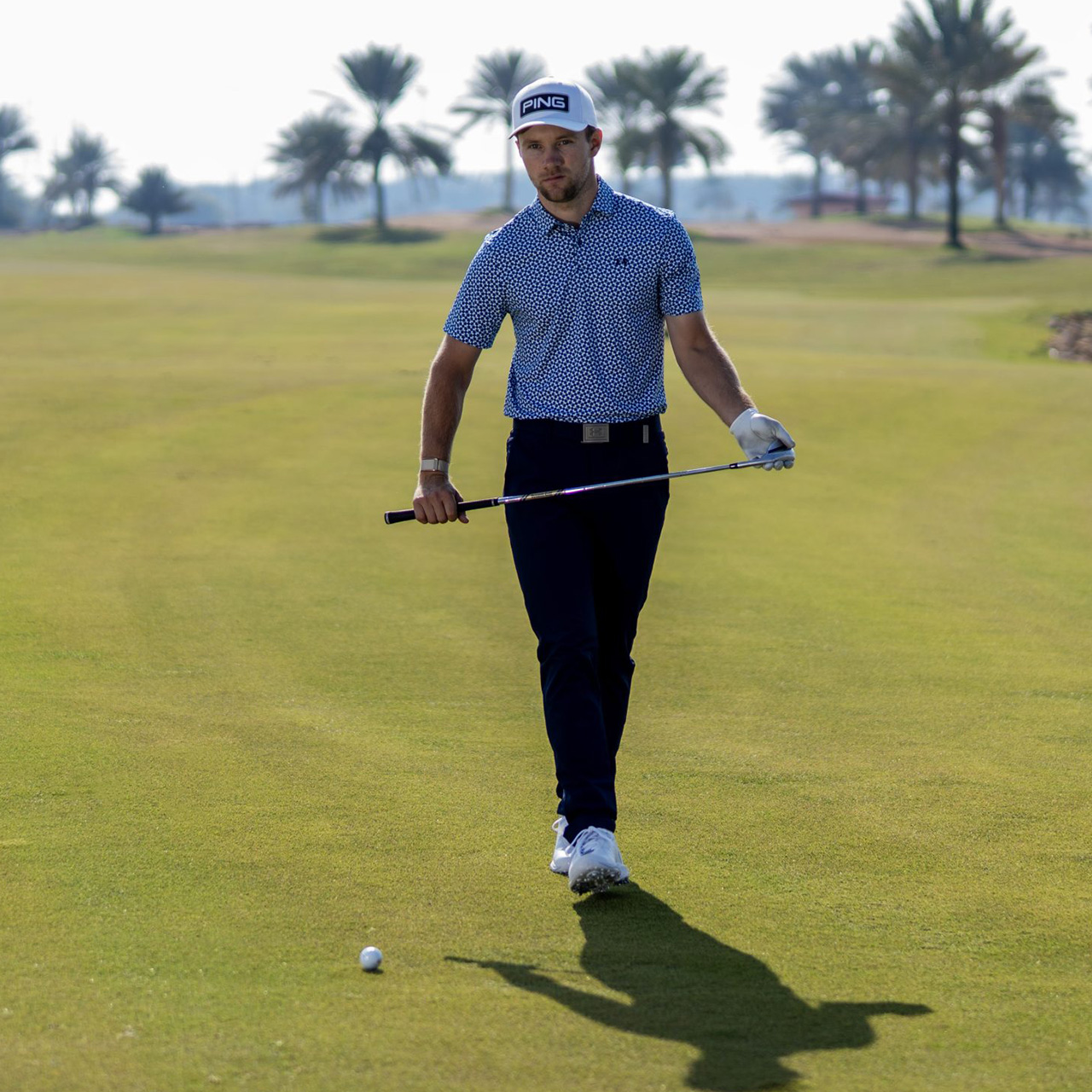 Male golfer on course wearing blue patterned polo shirt and navy trousers, holding golf club with palm trees in background