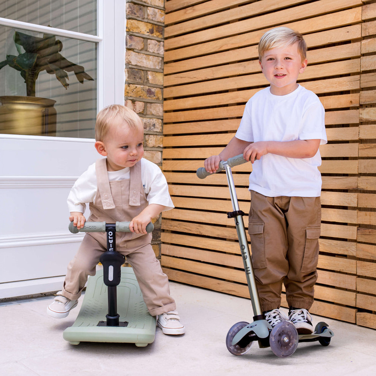 Two young children riding scooters outdoors on a paved patio with wooden and brick walls