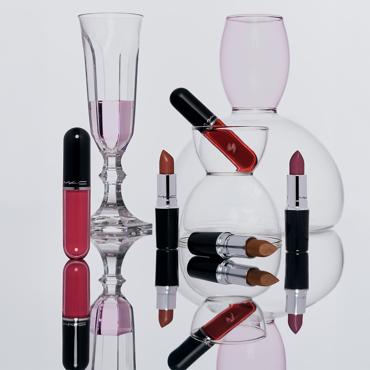 M.A.C Lustreglass lip range with lipsticks, nail polish, and glassware displayed on neutral background