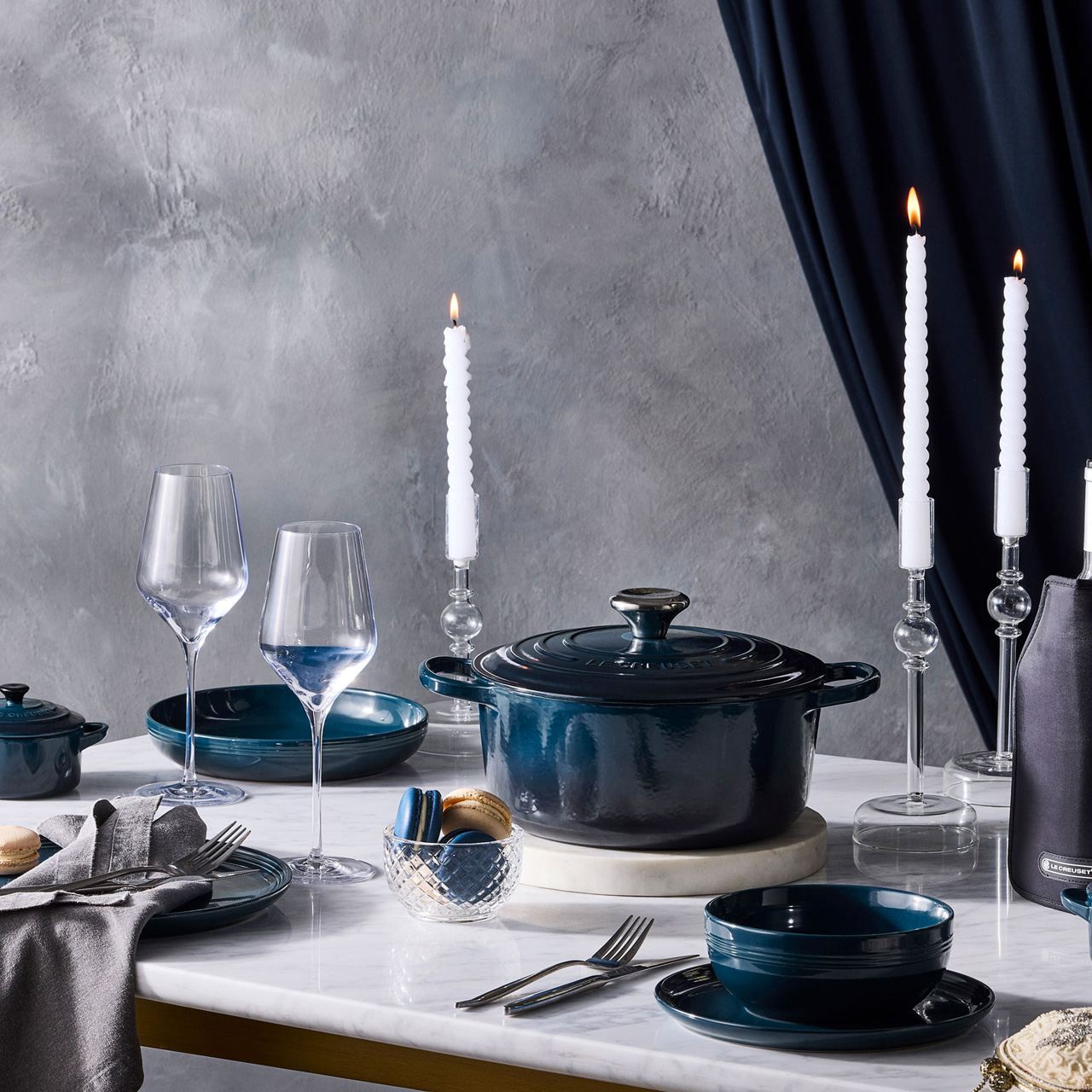Elegant table setting with dark blue cookware, wine glasses, white taper candles, and place settings against a grey textured backdrop