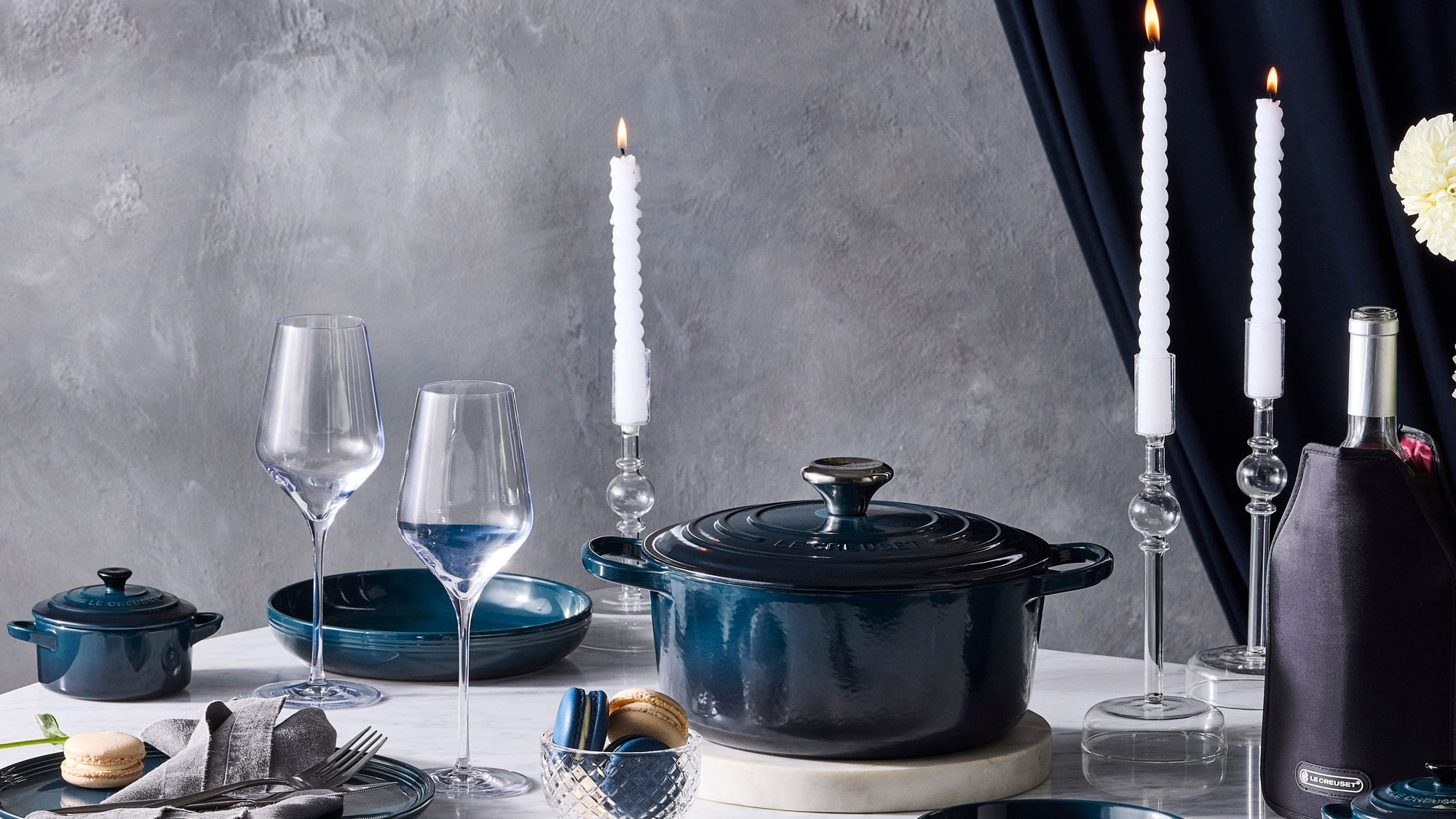 Elegant table setting with dark blue cookware, wine glasses, white taper candles, and place settings against a grey textured backdrop