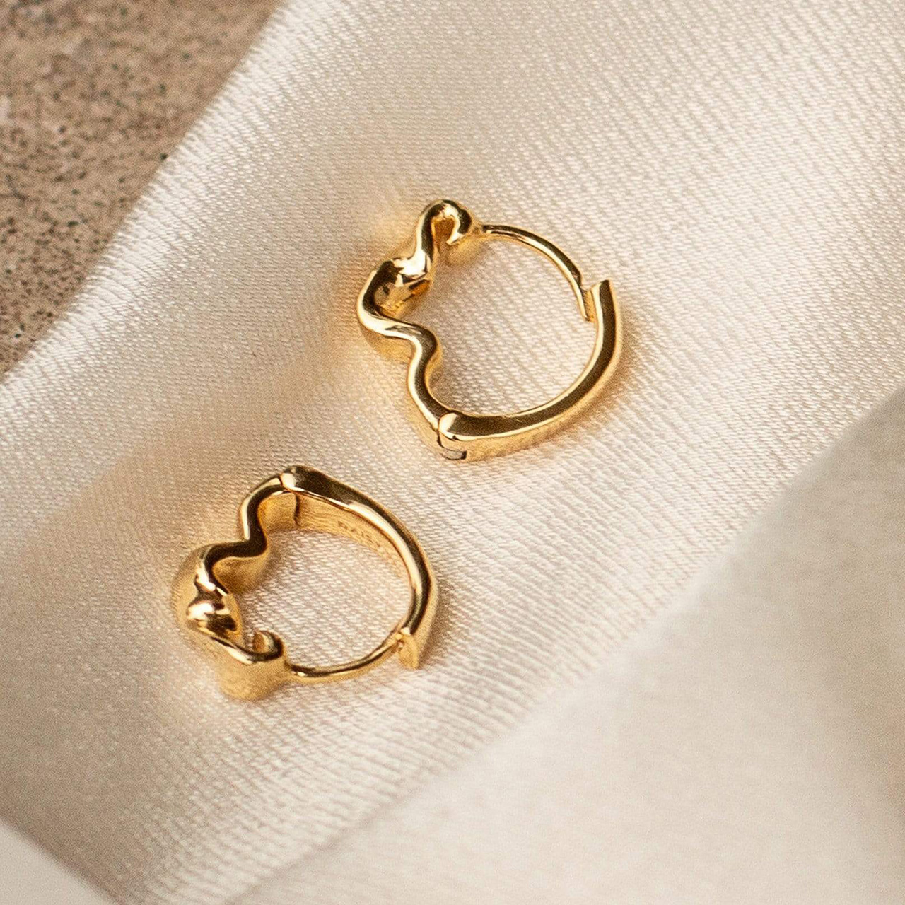 Two gold wavy hoop earrings on cream fabric with neutral backdrop