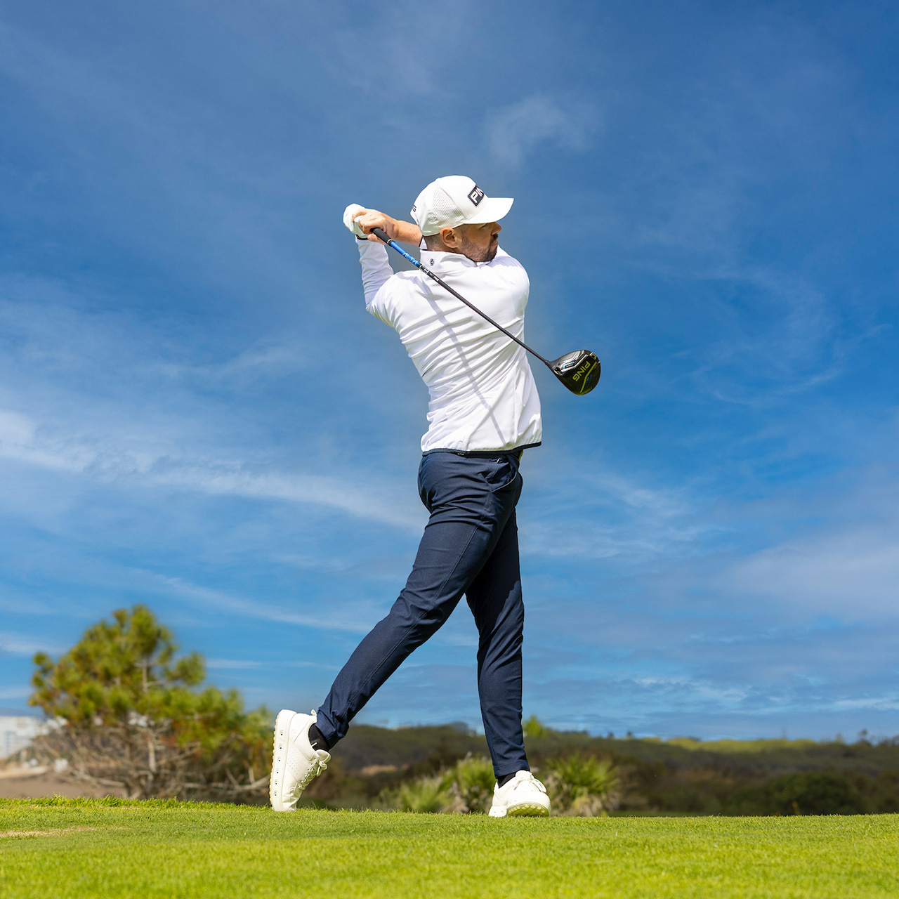 Golfer in white cap and navy trousers mid-swing on coastal golf course under blue sky