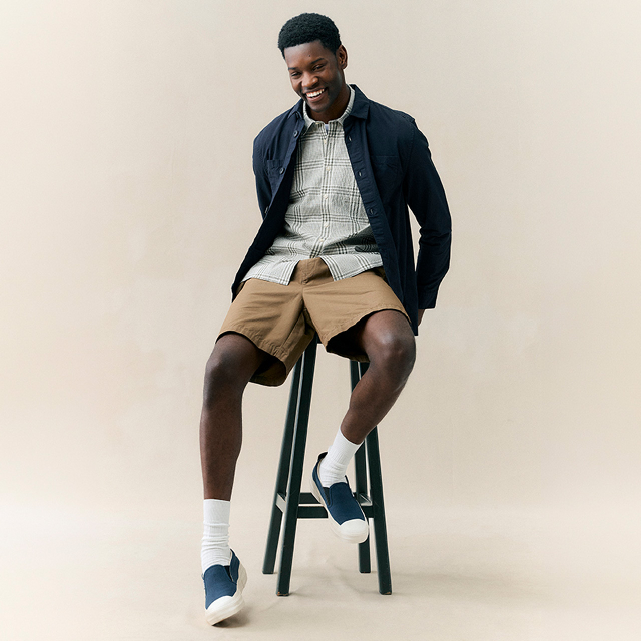 Man sitting on stool wearing navy jacket, checked shirt, and beige shorts with white socks