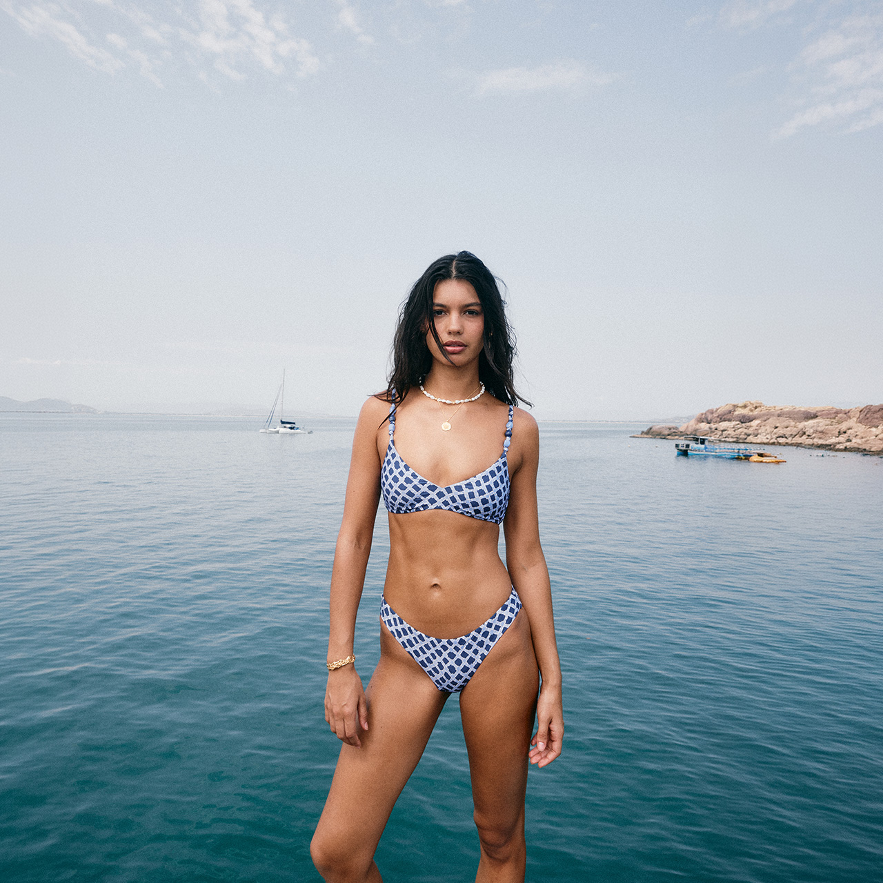 A woman in a checkered bikini stands by the sea with a rocky shoreline in the background under a clear sky.