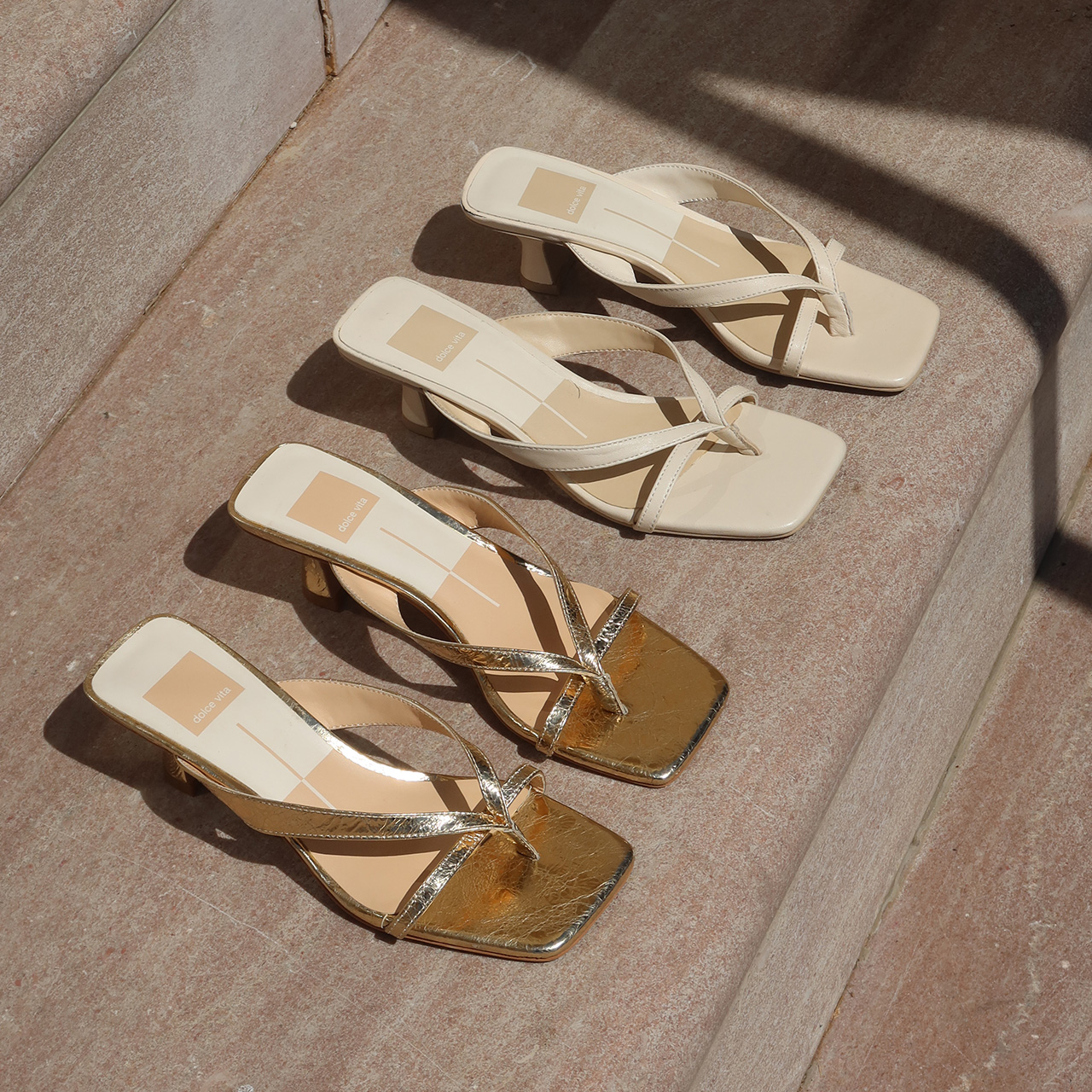 Four white and gold square-toe heeled sandals arranged on light stone floor