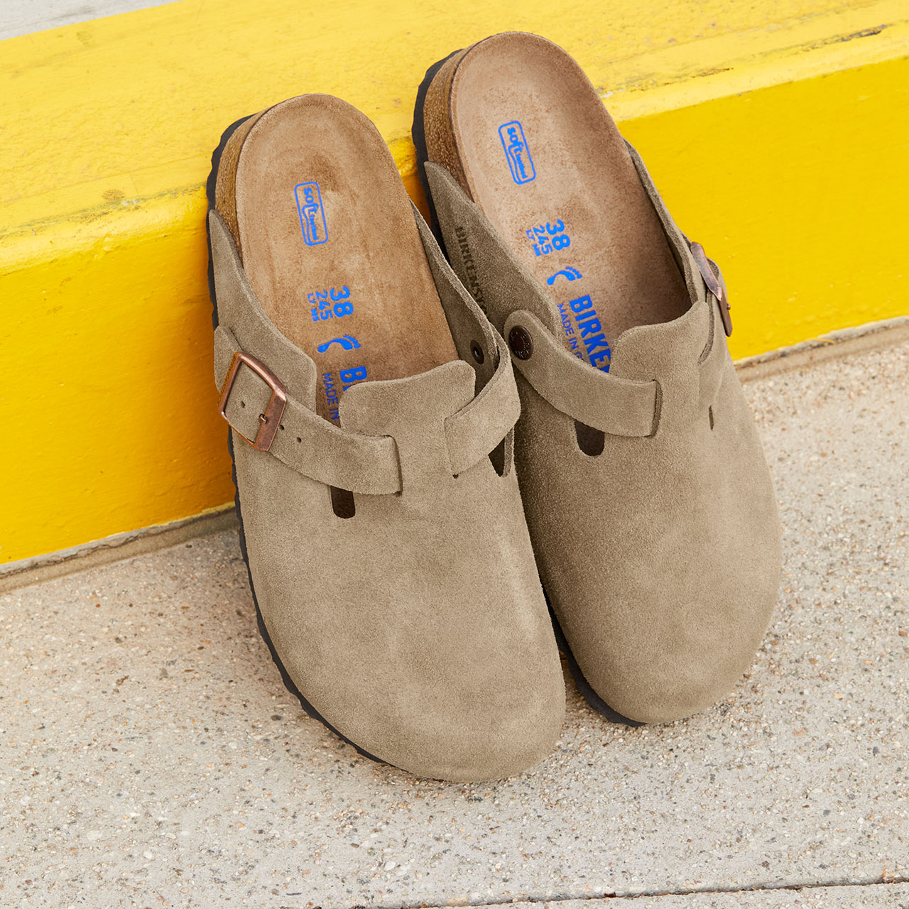 Pair of beige suede clogs with buckle detail and blue branded insoles against yellow wall