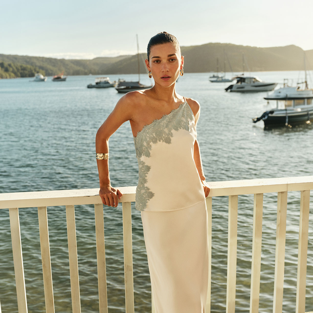 Woman wearing cream one-shoulder lace dress with gold jewellery, standing at waterfront railing with boats in background