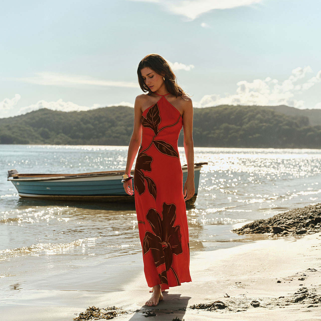 Woman in red floral halter dress standing on beach with sailing boat in background