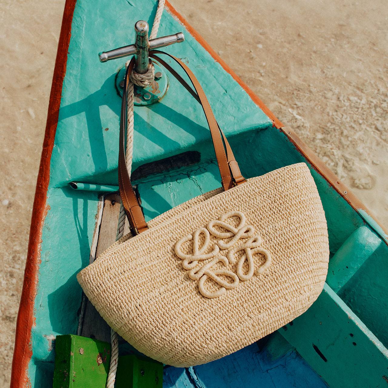 Natural woven tote bag with embroidered logo and leather handles resting on turquoise wooden boat