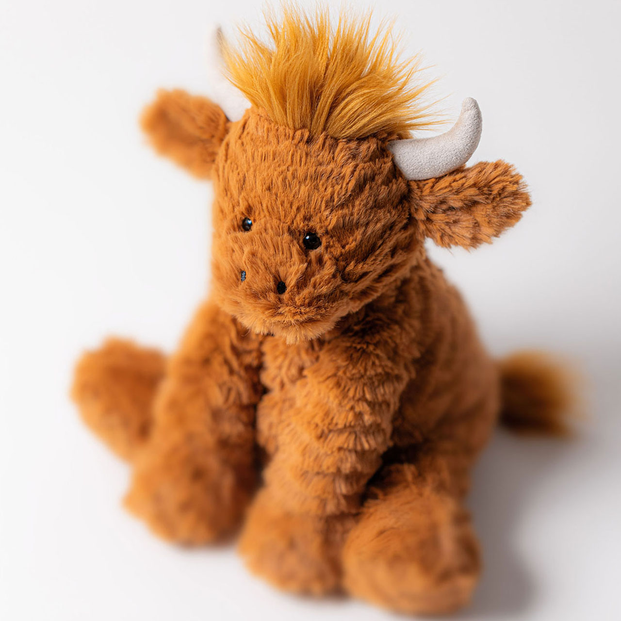 Soft golden-brown Jellycat plush toy Highland cow with horns and fluffy texture against white background