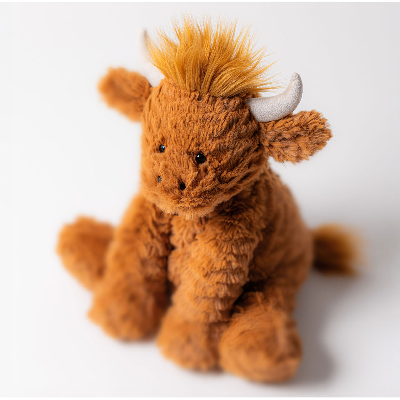 Soft golden-brown Jellycat plush toy Highland cow with horns and fluffy texture against white background