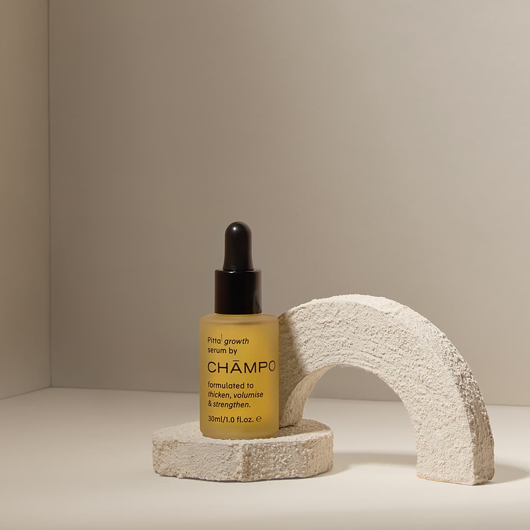 Yellow bottle of Champo hair serum with a dropper cap, placed on a textured stone pedestal. The pedestal is shaped like a curved arch, set in a corner with neutral lighting. The bottle label mentions growth, thickness, volume, and strength.
