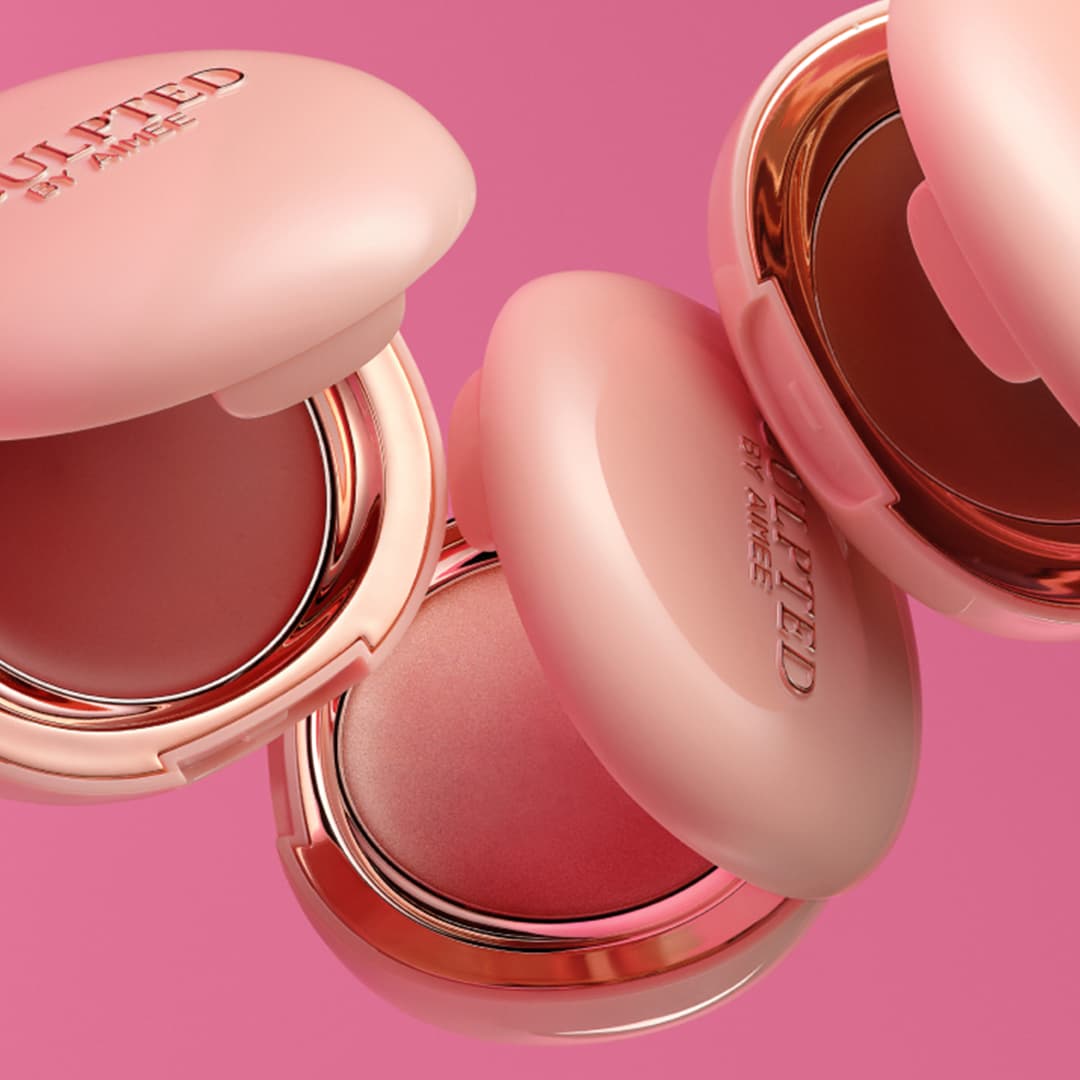 Pink and gold compact cases containing blush on a matching pink background. The cases are branded 'Sculpted by Aimee' and vary in shades of pink. Some are open, displaying the blush, while others are closed, highlighting the brand logo.