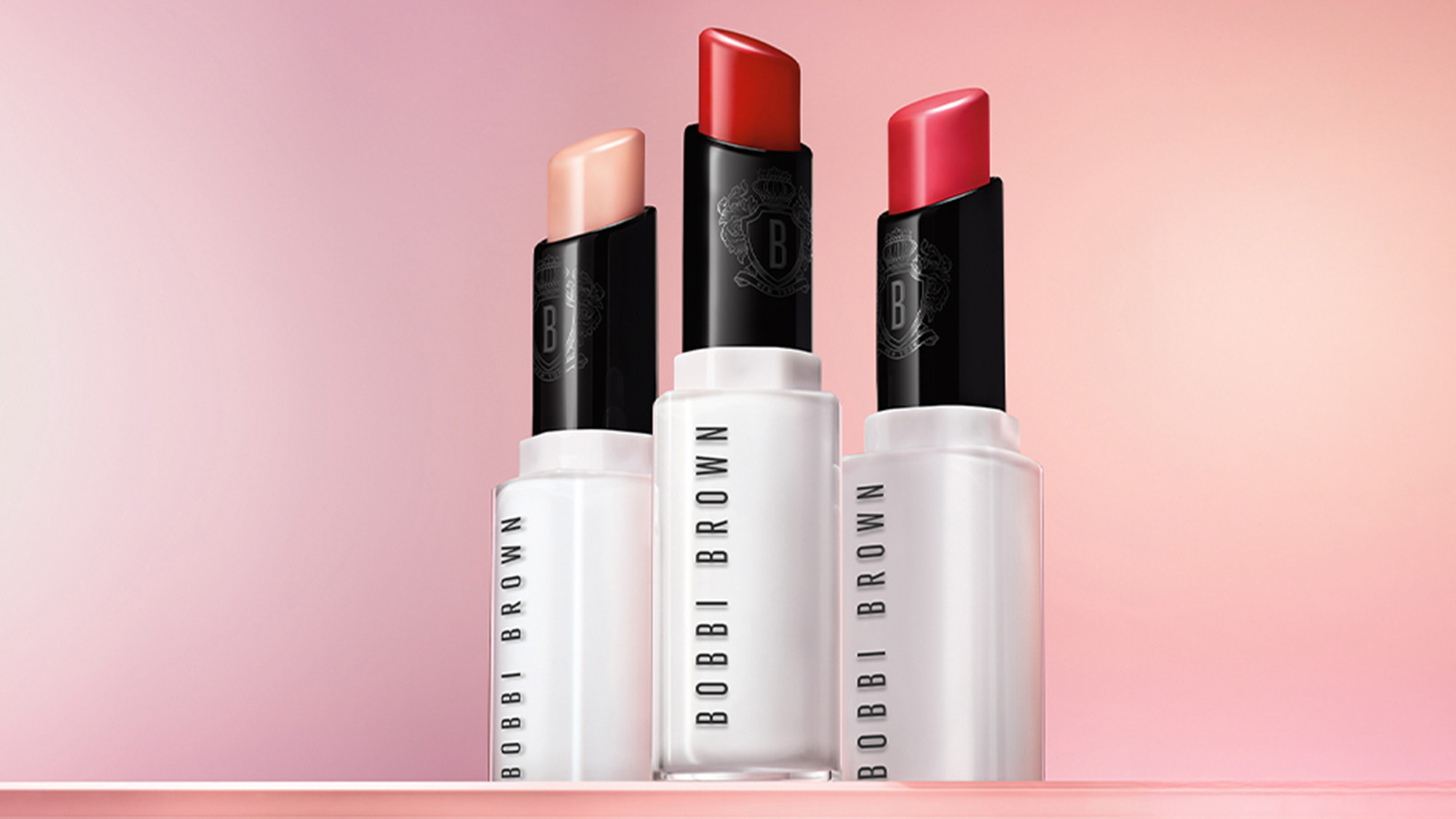 Three Bobbi Brown lipsticks in light, coral, and red shades against a pink gradient background.