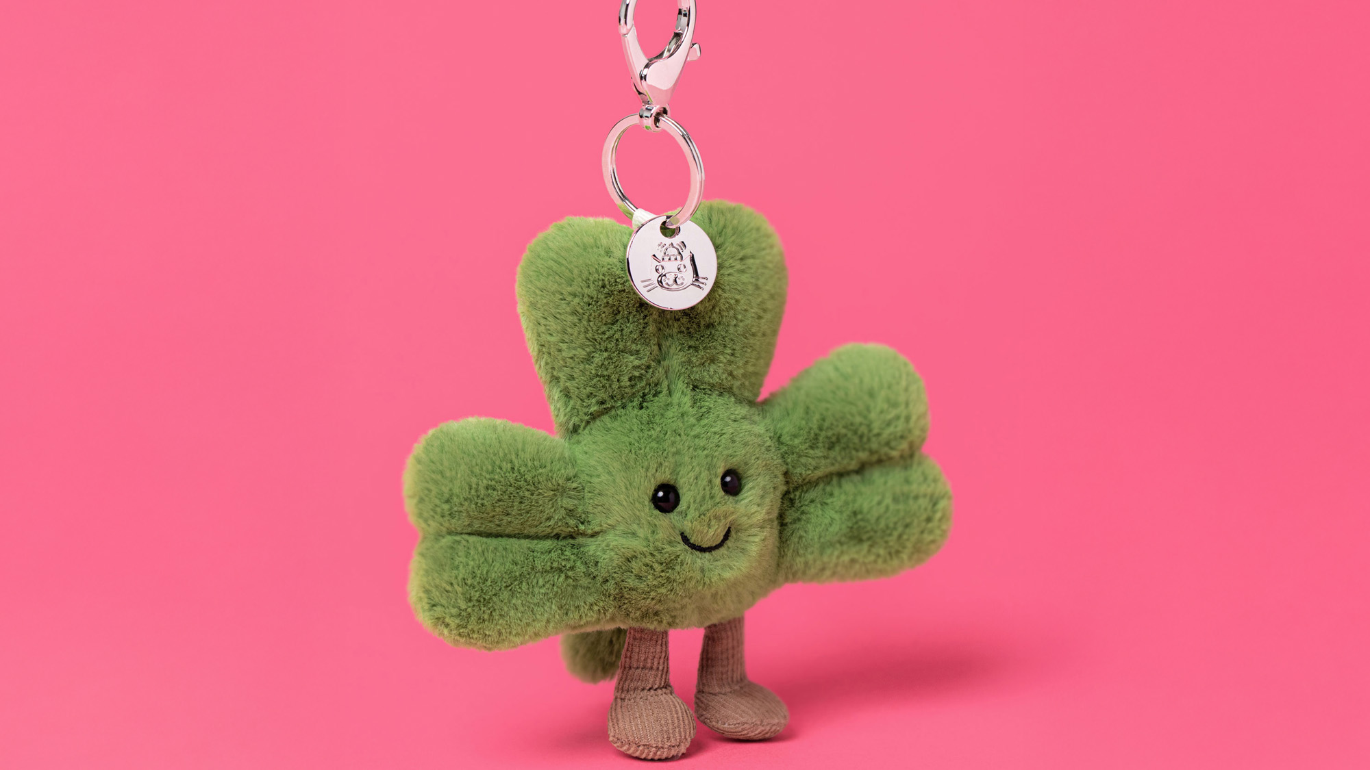 Plush green four-leaf clover keychain with a smiling face and small legs on a pink background.