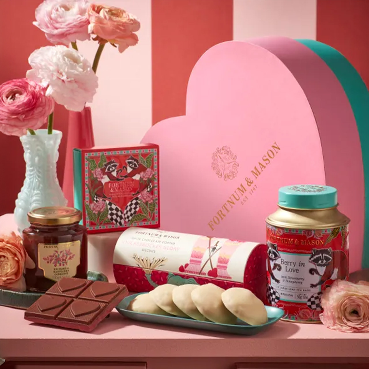 A heart-shaped box with pink flowers, tea, chocolate, and sweets arranged on a striped background.