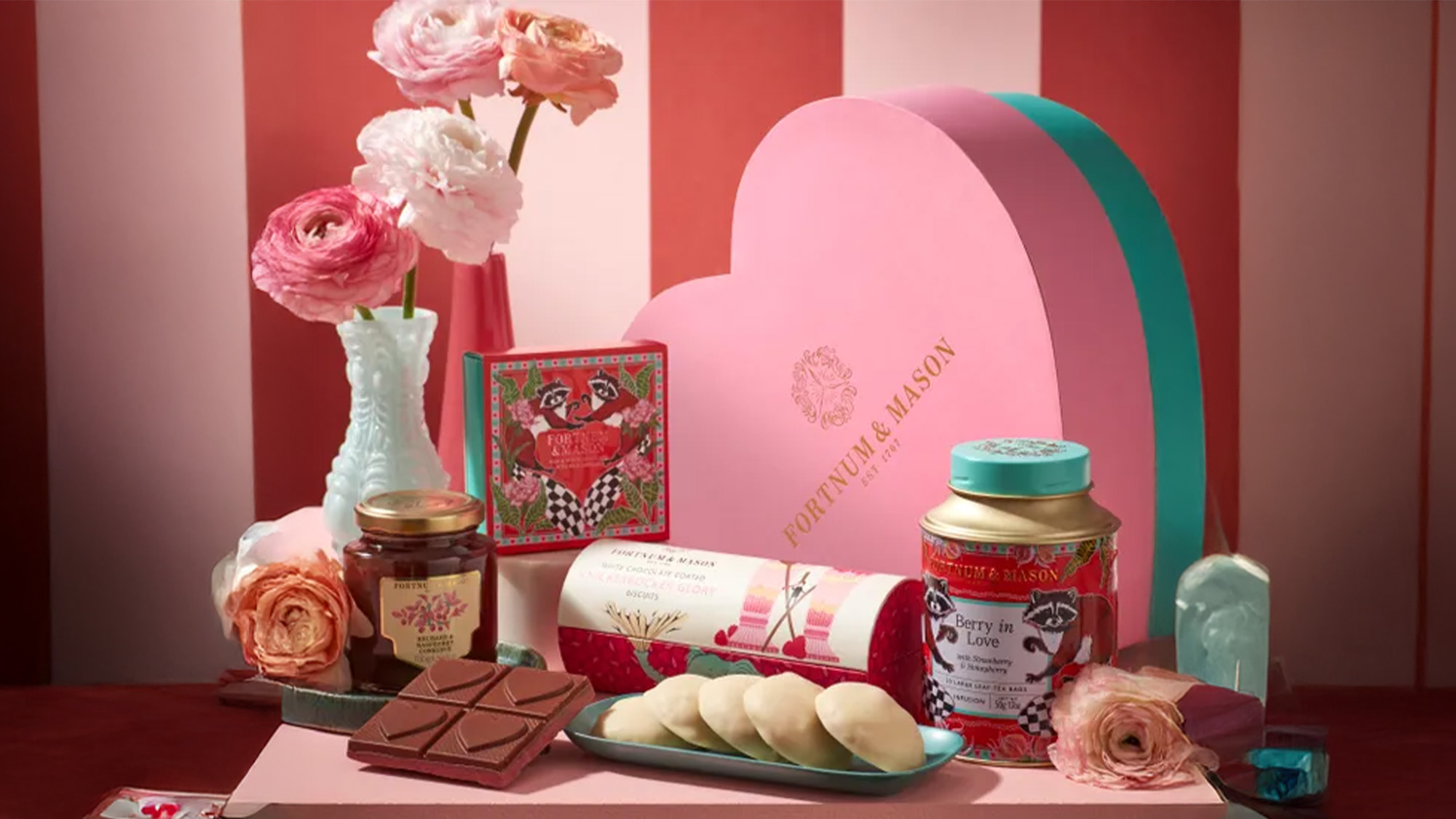 A heart-shaped box with pink flowers, tea, chocolate, and sweets arranged on a striped background.