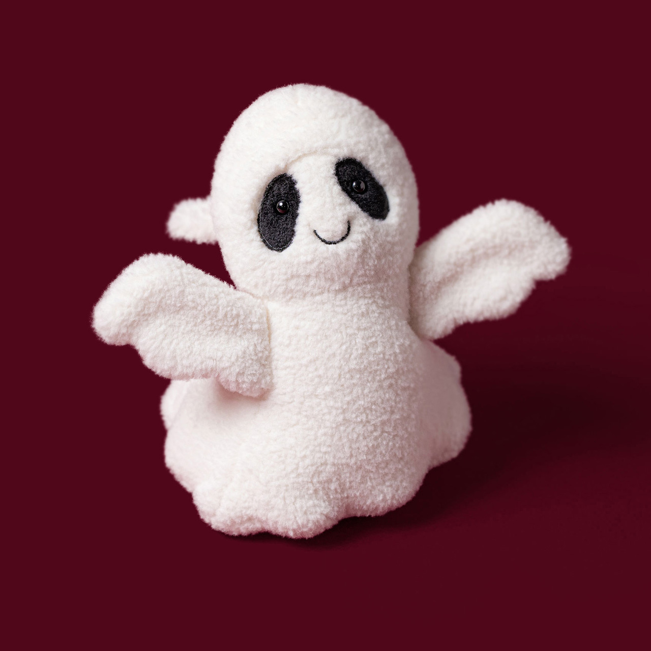 Cream-coloured fluffy Jellycat ghost toy with black eyes and smile on burgundy background