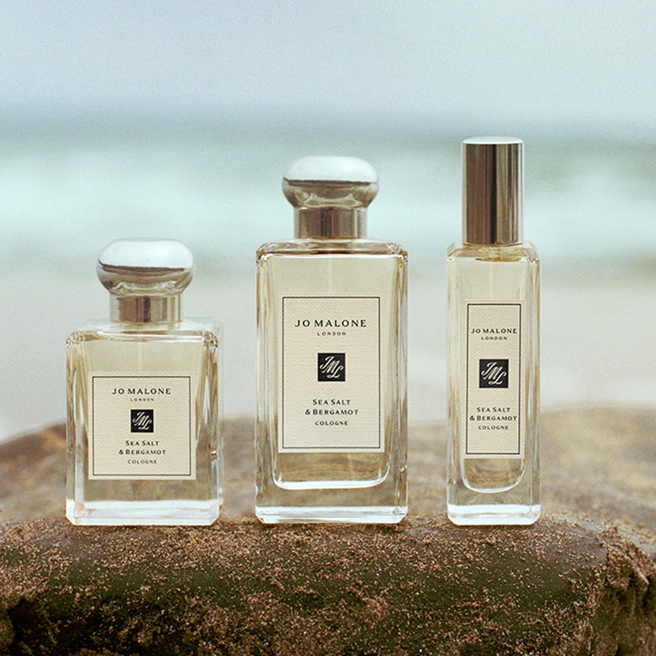 Three bottles of Jo Malone London Sea Salt & Bergamot cologne in varying sizes on sand with ocean background