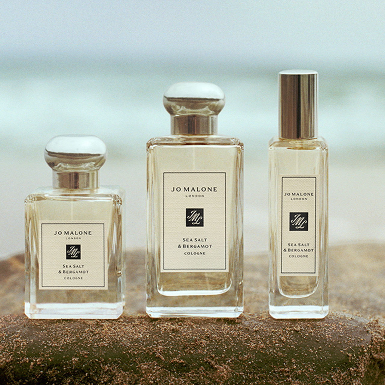 Three Jo Malone London Sea Salt & Bergamot cologne bottles in varying sizes arranged on sandy beach