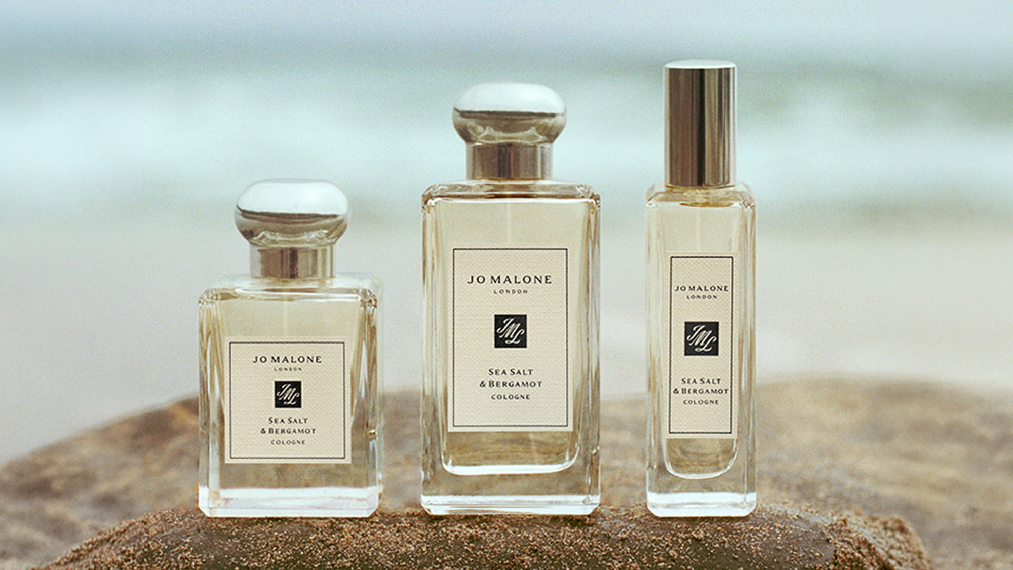 Three Jo Malone London Sea Salt & Bergamot cologne bottles in varying sizes arranged on sandy beach
