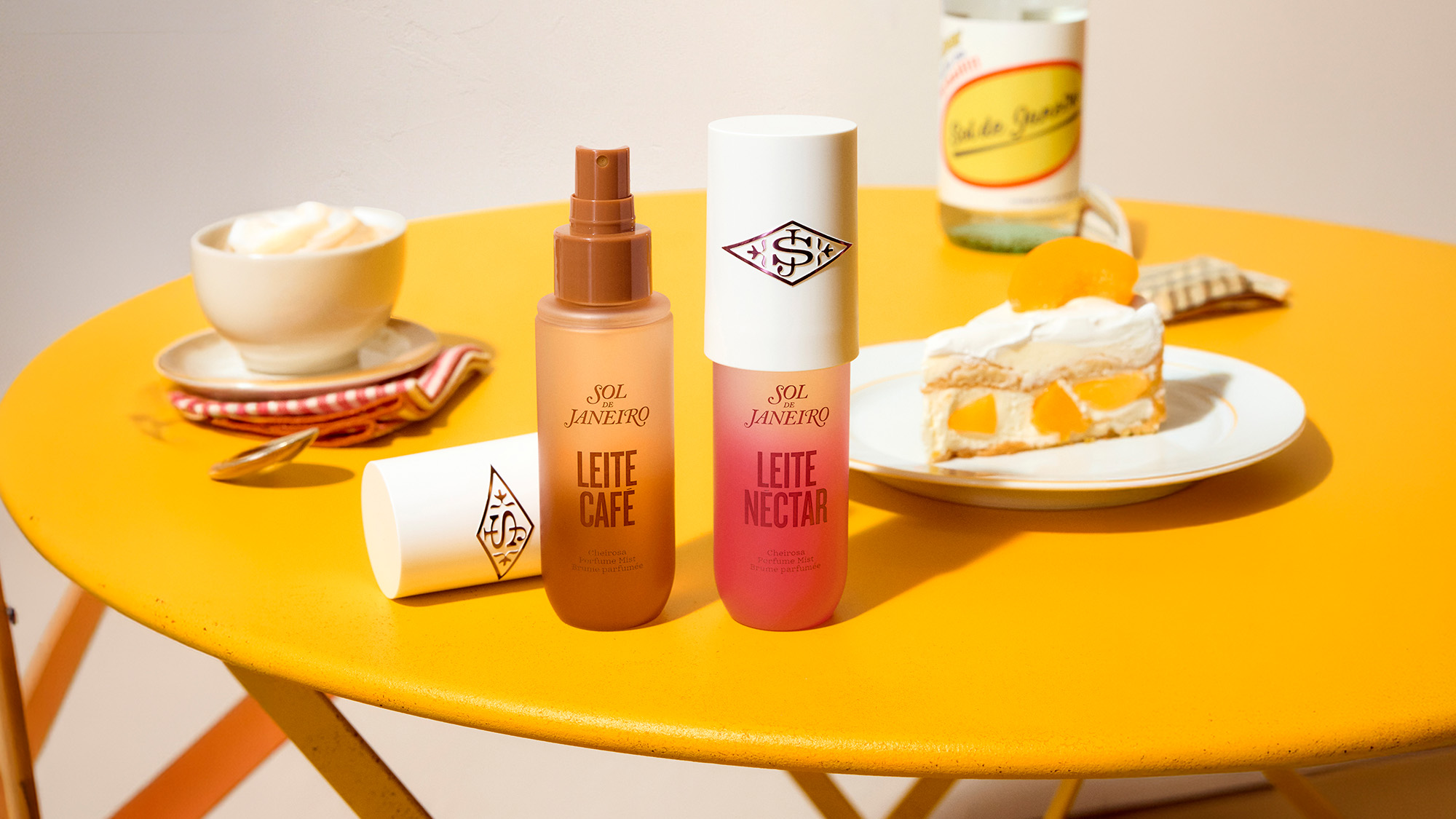Sol De Janeiro Leite perfume mists in latte and peach scents displayed on a yellow table with coffee and desserts