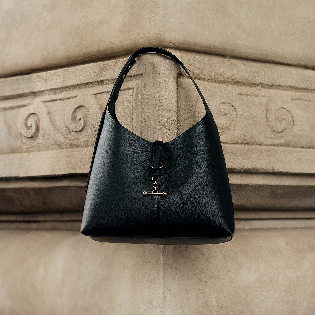 Black leather shoulder bag with toggle clasp detail displayed against ornate stone architectural backdrop