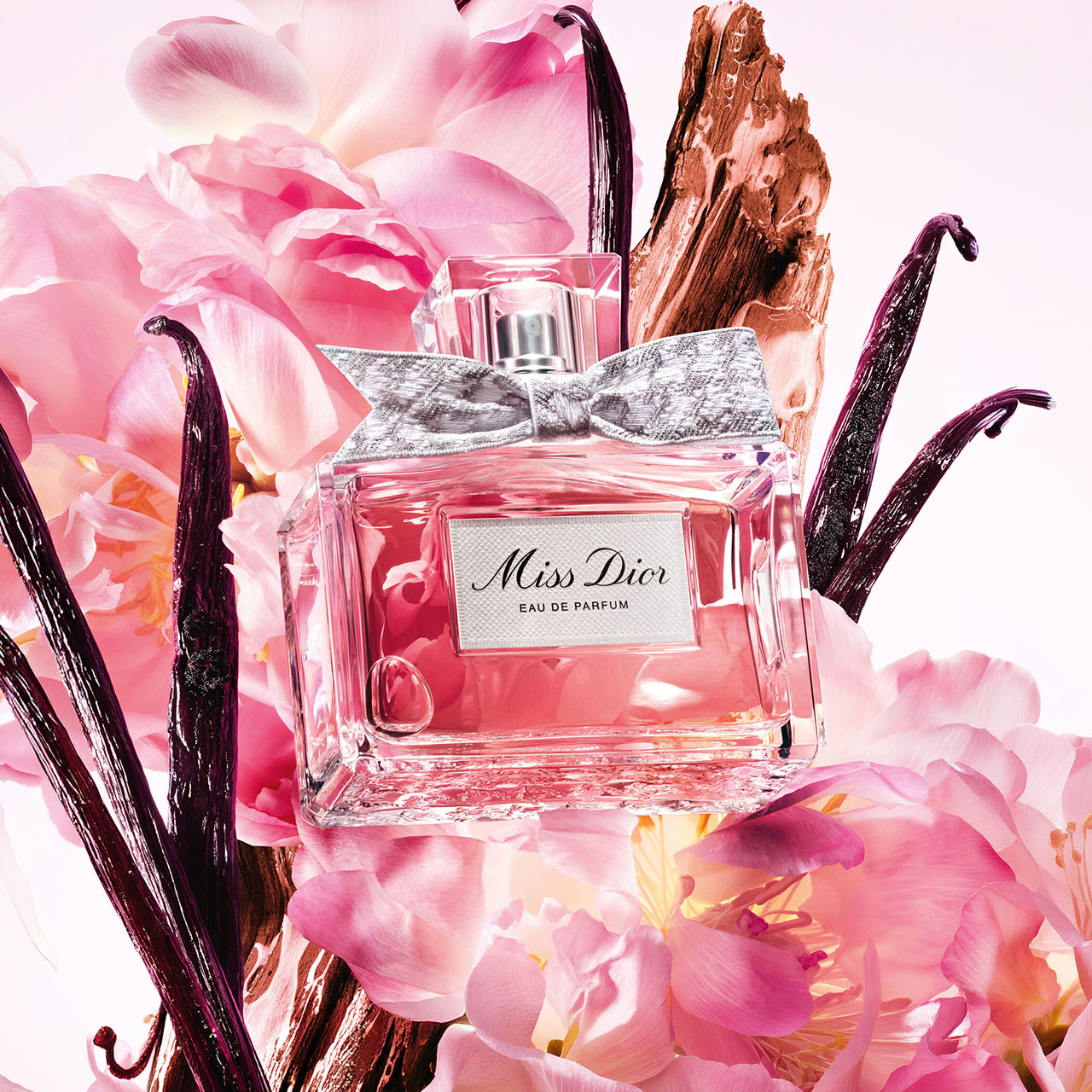 Miss Dior eau de parfum bottle with silver bow surrounded by pink peonies and dark vanilla pods on white background.