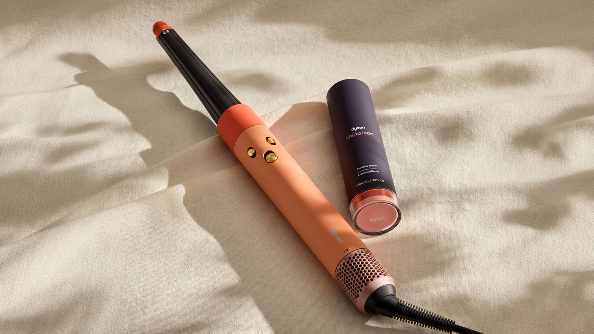 Dyson hair care styling tools including a curling iron and hair treatment product displayed on cream textured fabric
