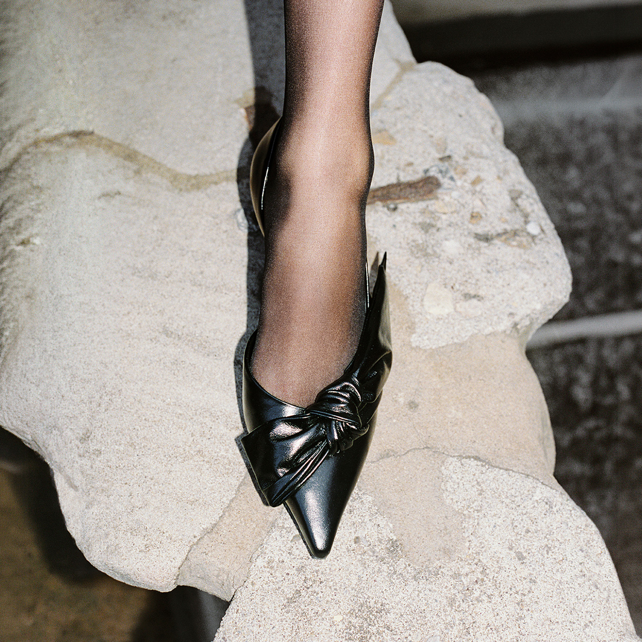 Woman wearing black pointed pump with gathered bow detail on foot, standing on stone steps