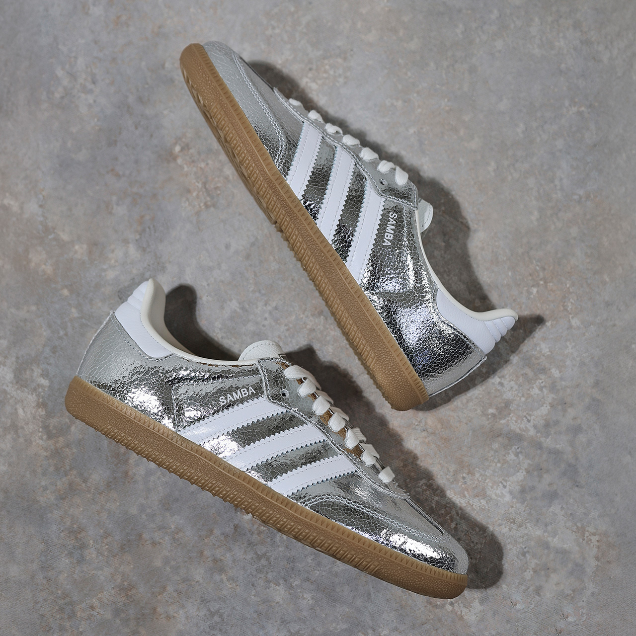 Silver metallic trainers with white stripes and gum soles are placed on a textured, grey surface. The brand name 'Samba' is visible on the side. The shoes have a sleek, retro design.