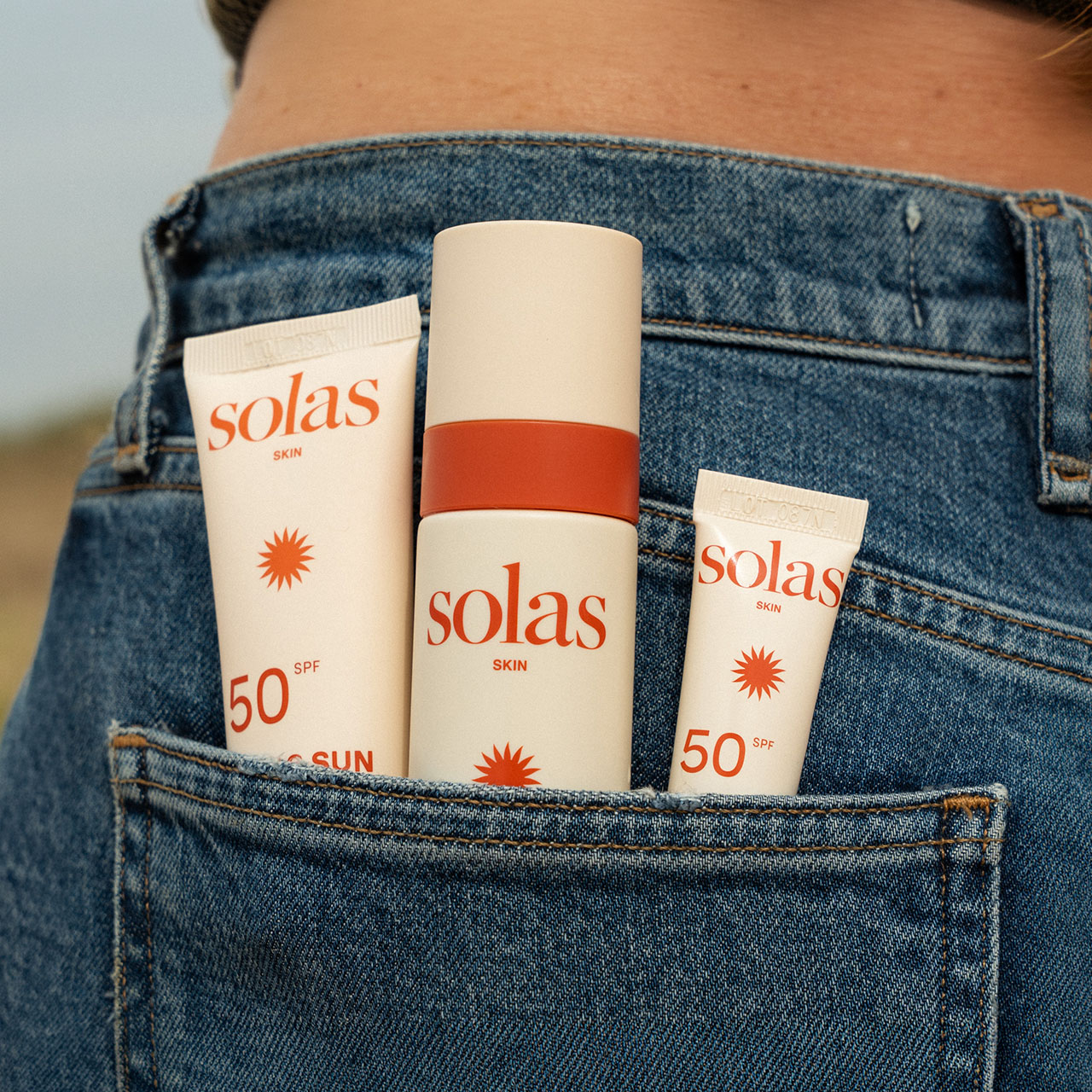 Three Solas Skin SPF 50 sunscreen products displayed in jeans back pocket with countryside background.