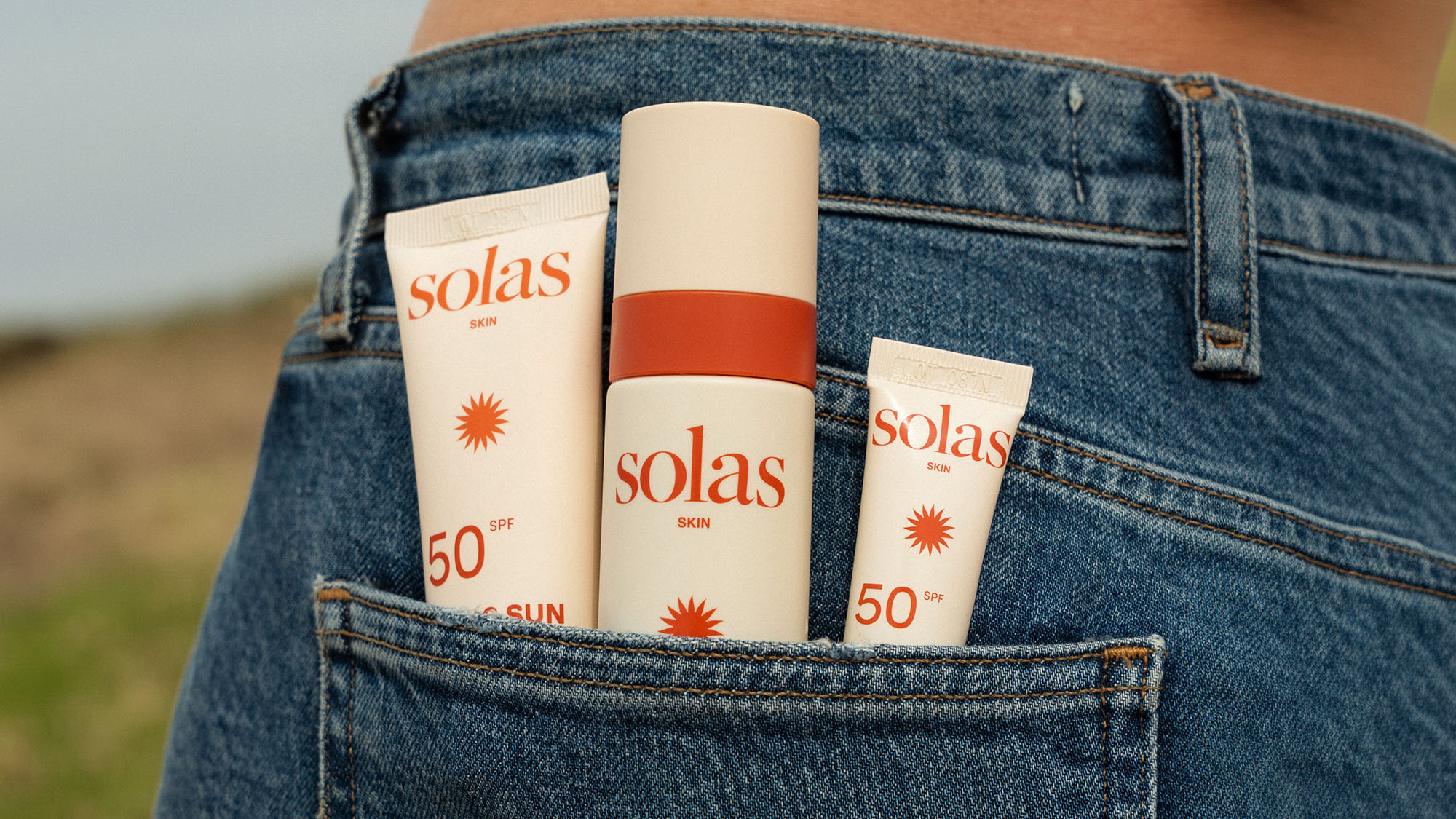 Three Solas Skin SPF 50 sunscreen products displayed in jeans back pocket with countryside background.
