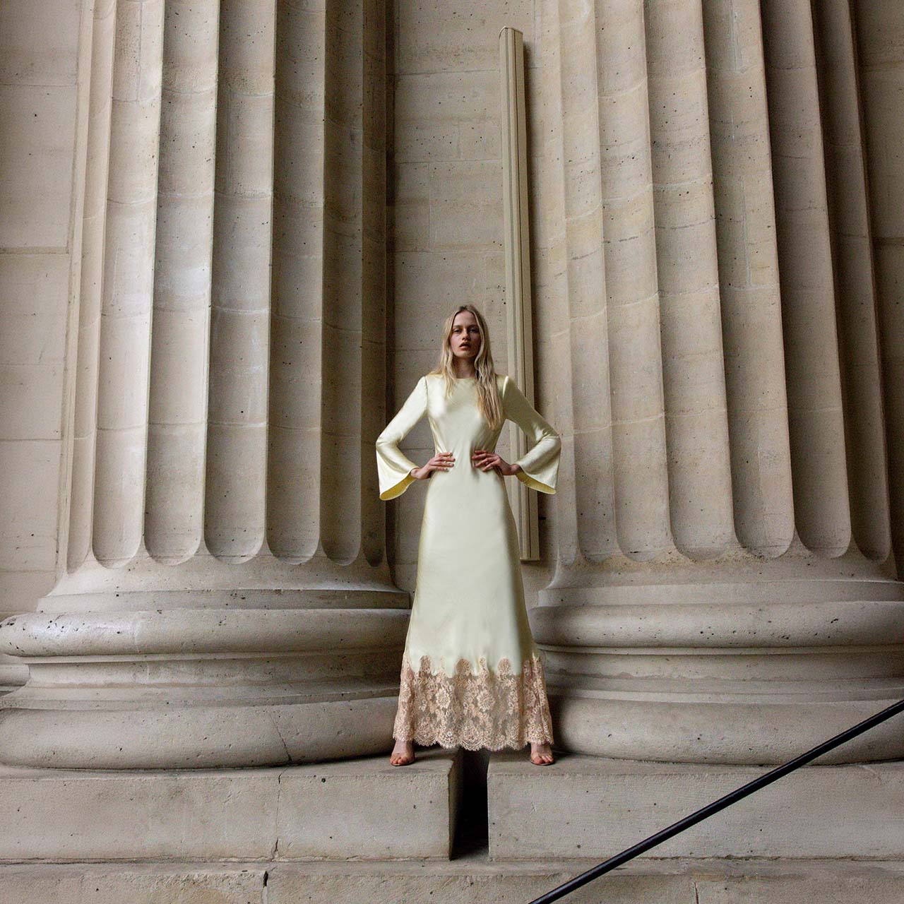 Woman wearing cream long-sleeved dress with lace hemline standing against architectural stone columns