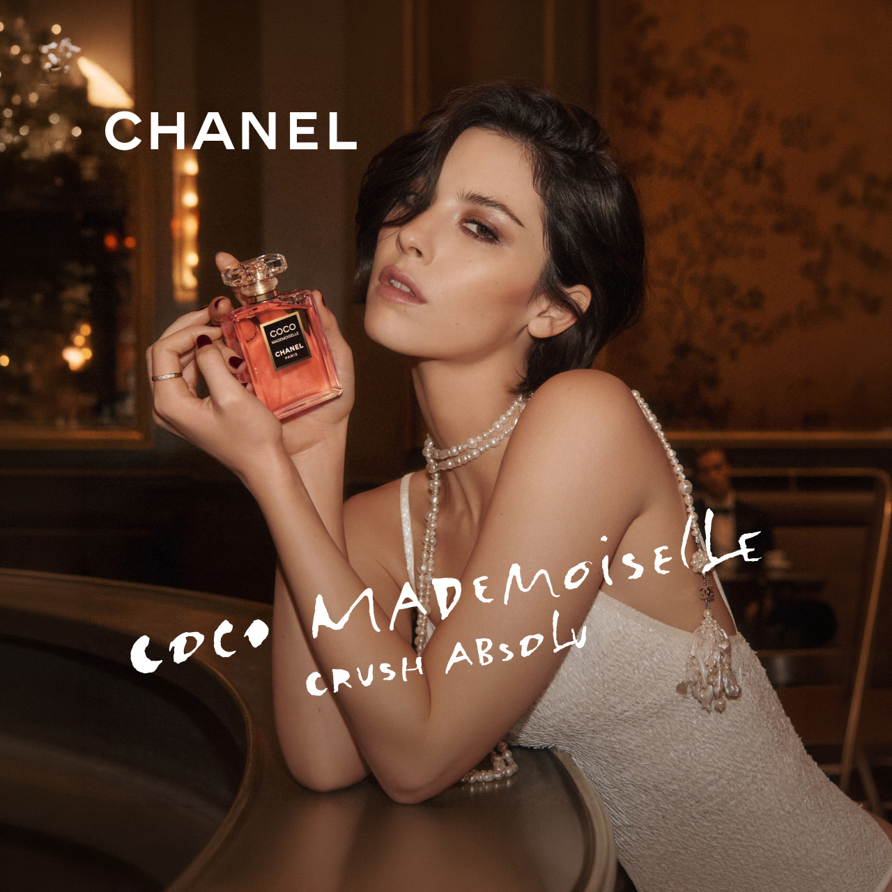 Woman holding red Chanel Coco Mademoiselle Crush Absolu fragrance bottle, wearing pearl jewellery against gold-lit interior backdrop