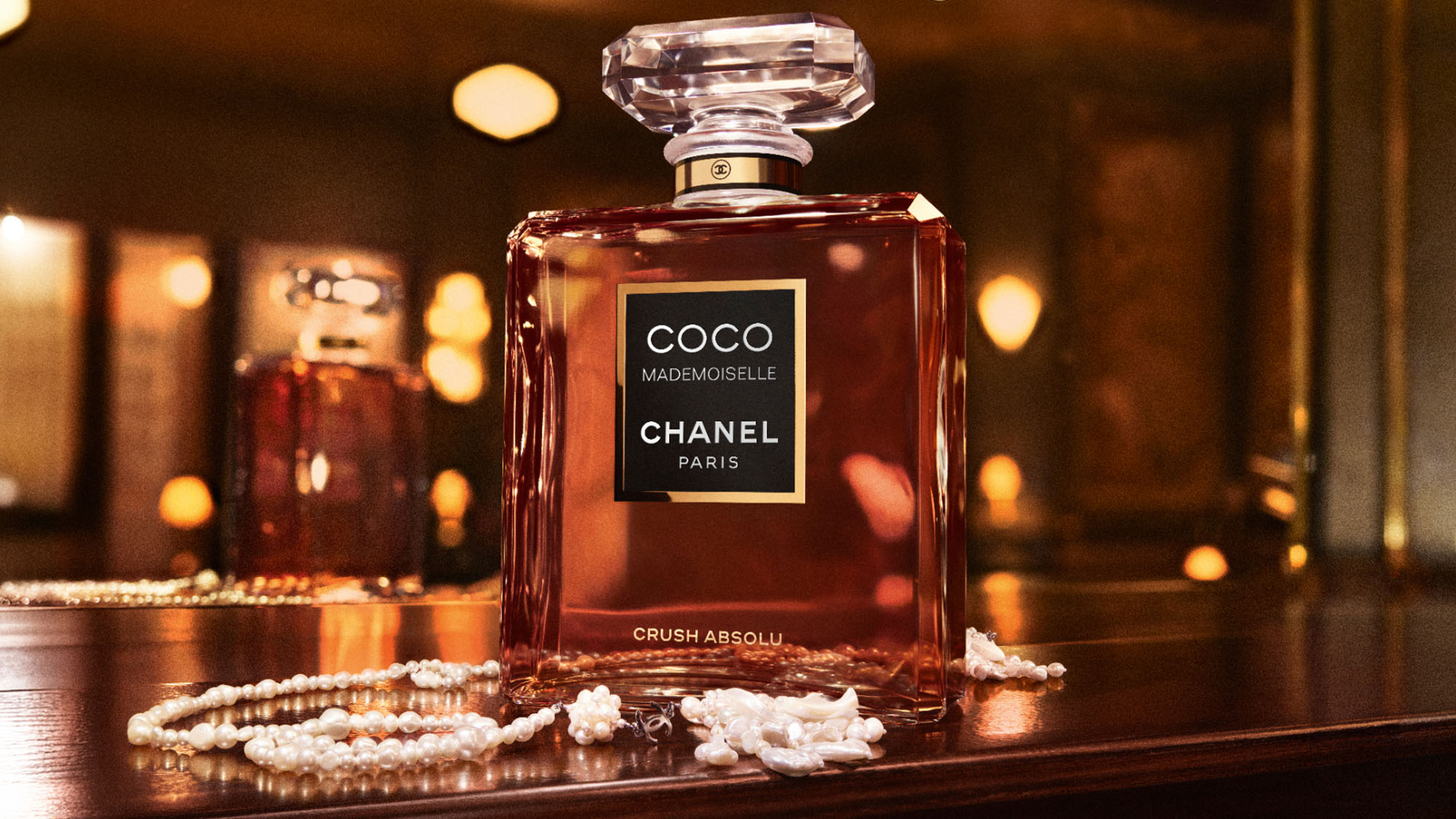Chanel Coco Mademoiselle Crush Absolu fragrance bottle with crystal stopper, surrounded by pearl jewellery on dark surface with warm lighting